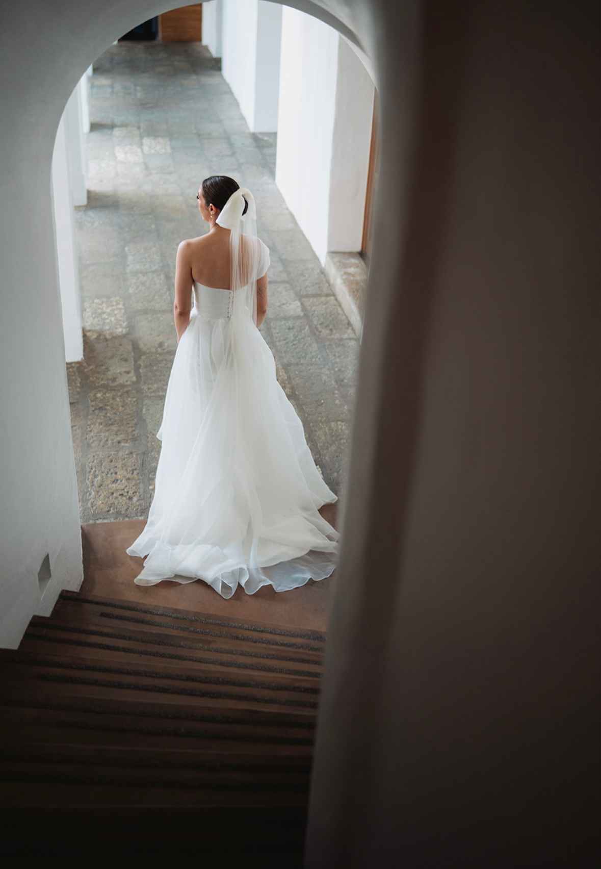 A bride in a white one-shoulder gown with a long veil, photographed from behind